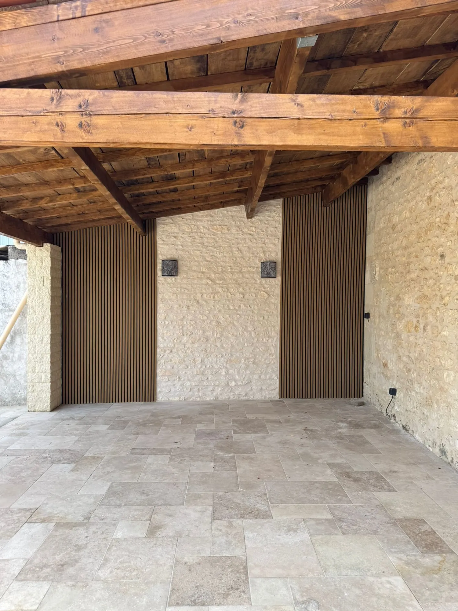 Sol en travertin posé en opus incertum sous un préau à charpente bois, contre un mur en pierre calcaire rejointoyé.